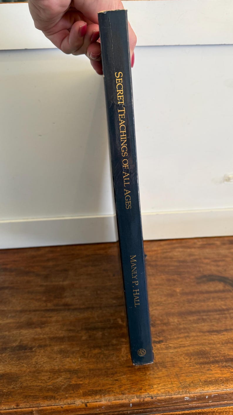 Może przedstawiać: Ciemnoniebieska książka zatytułowana "Secret Teachings of All Ages" autorstwa Manly P. Halla. Tytuł książki i nazwisko autora są w złotych literach. Książka jest trzymana pionowo.