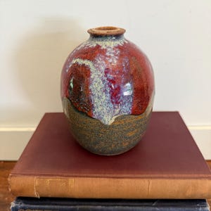 May include: A handcrafted ceramic vase with a rounded body and a small opening. The vase features a rich color palette, with a deep red upper portion, transitioning to a speckled, textured brown base. The vase is displayed on a stack of vintage books.