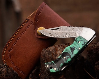 May include: A green and black patterned pocket knife with a damascus steel blade, open and resting on a brown leather sheath. The knife is resting on a piece of wood.
