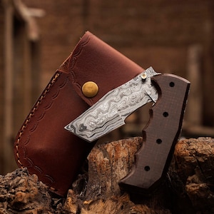 Puede incluir: Una navaja plegable con una hoja de acero estampada y un mango de madera oscura. El cuchillo se combina con una funda de cuero marrón con un borde decorativo y un cierre de botón de latón. El cuchillo y la funda se exhiben sobre un trozo de madera.