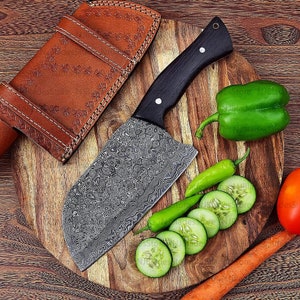 May include: A large, black-handled cleaver with a patterned, stainless steel blade sits on a wooden cutting board. The cleaver is next to a leather sheath, a green bell pepper, a red tomato, green chili peppers, and sliced cucumbers.