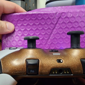 May include: A custom video game controller with a bronze faceplate and black joysticks. The controller has white and black accents, with the letters "Fn" on the buttons. A purple bubble mailer is in the background.