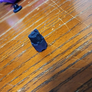 May include: A small, black, 3D-printed object with a complex, geometric design. The object is resting on a wooden surface with a visible grain pattern. Another smaller black object is in the background.