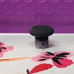 May include: A black joystick thumbstick with a textured surface. The thumbstick is set against a floral patterned background with pink and red flowers. The background is a purple, textured surface.