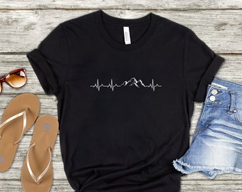 Könnte beinhalten: Schwarzes T-Shirt mit einem weißen Bergketten-Motiv und einer Herzschlaglinie. Das Motiv ist mittig auf dem T-Shirt platziert. Das T-Shirt liegt flach auf einer Holzoberfläche.