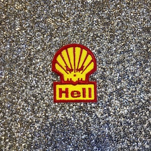 Może przedstawiać: Żółto-czerwona naszywka z haftem przedstawiająca stylizowany wzór muszli z napisem "Hell" w żółtych literach. Naszywka znajduje się na błyszczącym, srebrnym tle.