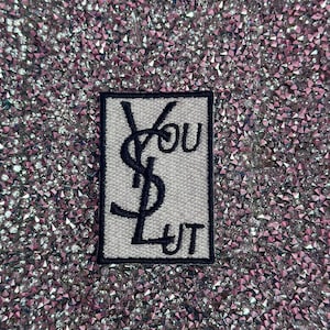 May include: A white patch with black embroidery of the Yves Saint Laurent logo and the text "YOU JUST" on a sparkly pink background.