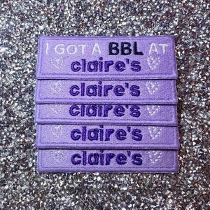 Op de afbeelding: Vijf paarse stoffen patches met witte stiksels. De bovenste patch heeft de tekst "I GOT A BBL AT claire's". De overige vier patches hebben de tekst "claire's" met een klein wit hartontwerp.