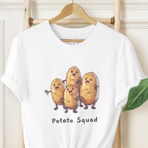 Pode incluir: Camiseta branca com um gráfico de quatro batatas de desenho animado com rostos sorridentes. As batatas estão de mãos dadas e o texto "Potato Squad" está impresso abaixo.