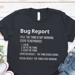 Peut inclure: Un t-shirt bleu foncé avec du texte blanc qui dit "Bug Report" et comprend une description humoristique d'un rapport de bug. Le t-shirt est posé à plat sur une surface en bois avec une paire de baskets blanches et un jean bleu.