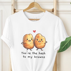 Puede incluir: Camiseta blanca con una linda ilustración de dos patatas tomándose de la mano. El texto "You're the hash to my browns" está impreso en la camiseta.