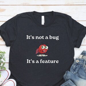 以下が含まれることがあります： 赤い漫画の虫の白いグラフィックが描かれた濃い青色のTシャツ。Tシャツには「バグではありません。機能です。」というテキストが印刷されています。