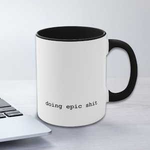 May include: White ceramic coffee mug with a black rim and handle. The mug has the text "doing epic shit" printed on the front in a typewriter font.