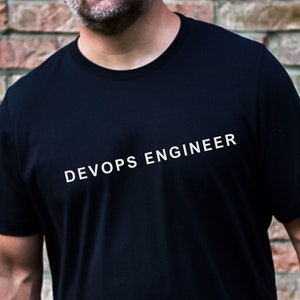 Peut inclure: T-shirt noir avec l'inscription blanche "DEVOPS ENGINEER" en diagonale sur la poitrine. Le t-shirt à manches courtes semble être fait d'une matière douce. En arrière-plan, un mur de briques.