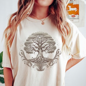 May include: A cream-colored oversized t-shirt with a brown tree of life graphic. The tree has intricate roots and branches, and is surrounded by a decorative floral design.