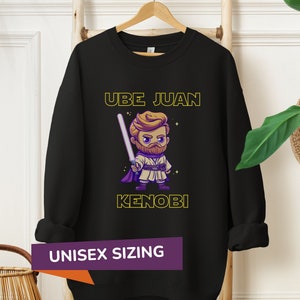 May include: Black sweatshirt with a cartoon image of a character holding a lightsaber. The text "UBE JUAN KENOBI" is printed in yellow on the shirt. The text "UNISEX SIZING" is printed on a purple tag.