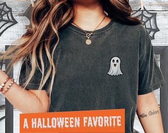 May include: A gray t-shirt with a cartoon ghost graphic and the text "A HALLOWEEN FAVORITE" printed on the front. The shirt is worn by a person with long brown hair and a gold necklace.