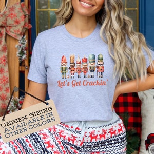 May include: A grey t-shirt featuring a watercolour illustration of a line of nutcracker figurines. The text "Let's Get Crackin'" is printed below the figurines. The t-shirt is available in other colours.