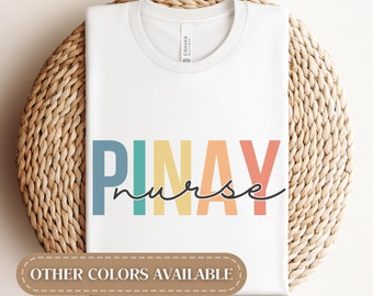 May include: White t-shirt with the text "PINAY nurse" in rainbow colors. The text is in a bold, blocky font. The words "OTHER COLORS AVAILABLE" are printed in brown at the bottom of the shirt.