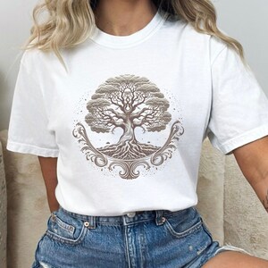 May include: White t-shirt with a brown tree of life design. The tree has intricate roots and branches, and is surrounded by a decorative border.