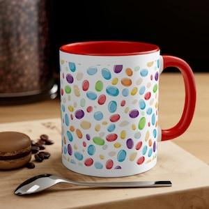 May include: White ceramic mug with a red interior and handle. The exterior is covered in a pattern of colorful jelly bean shapes. The mug is sitting on a wooden surface with a spoon, coffee beans, and a pastry.