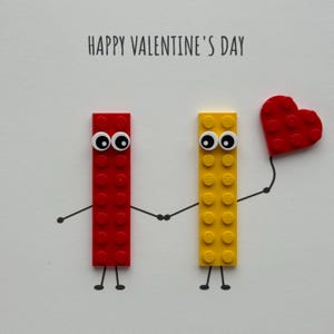 May include: A Valentine's Day card featuring two Lego brick figures holding hands. One red brick figure holds a red heart. The card reads "HAPPY VALENTINE'S DAY" in black text on a white background.