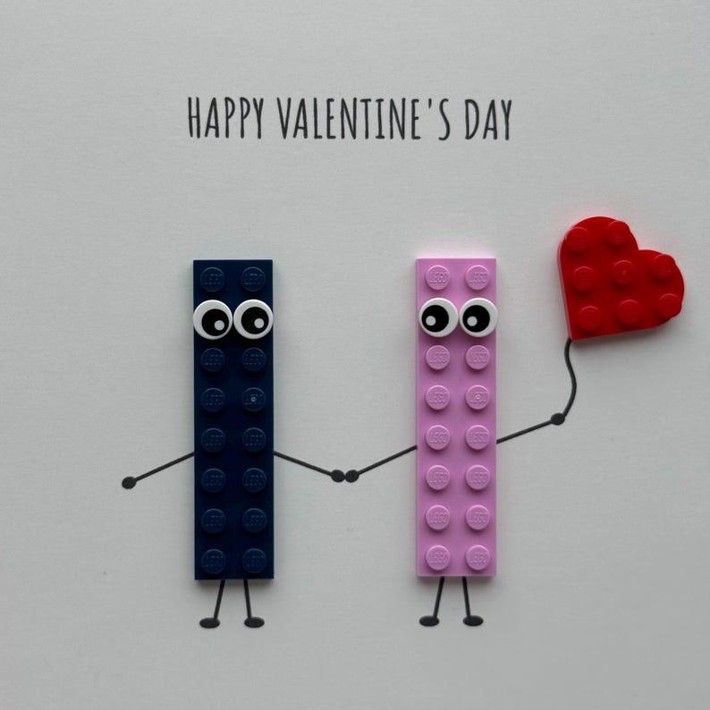 May include: A Valentine's Day card featuring two Lego brick figures with googly eyes holding hands. One figure is navy blue, the other is pink, and a red heart is held by the pink figure. The words "HAPPY VALENTINE'S DAY" are printed at the top.