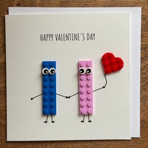 May include: Valentine's Day card with the text "HAPPY VALENTINE'S DAY". The card features two Lego figures, one blue and one pink, holding hands. A red Lego heart is held by the pink figure.