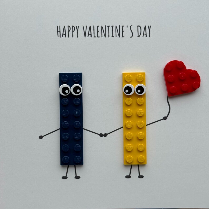 May include: A Valentine's Day card featuring two Lego brick figures holding hands. One figure is navy blue, the other is yellow, and both have googly eyes. A red heart is held by the yellow figure. The words "HAPPY VALENTINE'S DAY" are printed at the top.