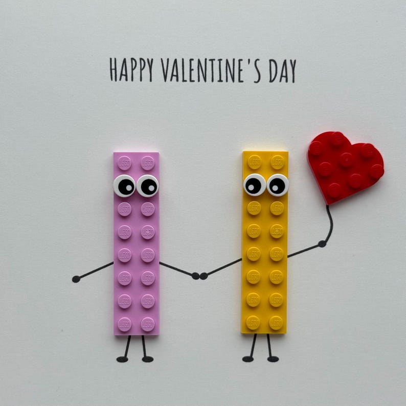 May include: A Valentine's Day card featuring two toy bricks with googly eyes holding hands. One brick is pink, the other yellow, and a red heart is held by the yellow brick. The words "HAPPY VALENTINE'S DAY" are printed at the top.