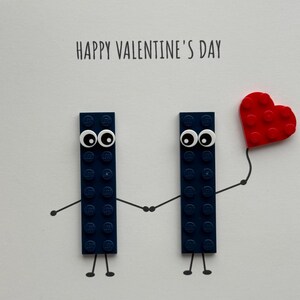 May include: A Valentine's Day card featuring two navy blue building blocks with googly eyes, holding hands, and a red heart balloon. The text "HAPPY VALENTINE'S DAY" is printed at the top of the card.