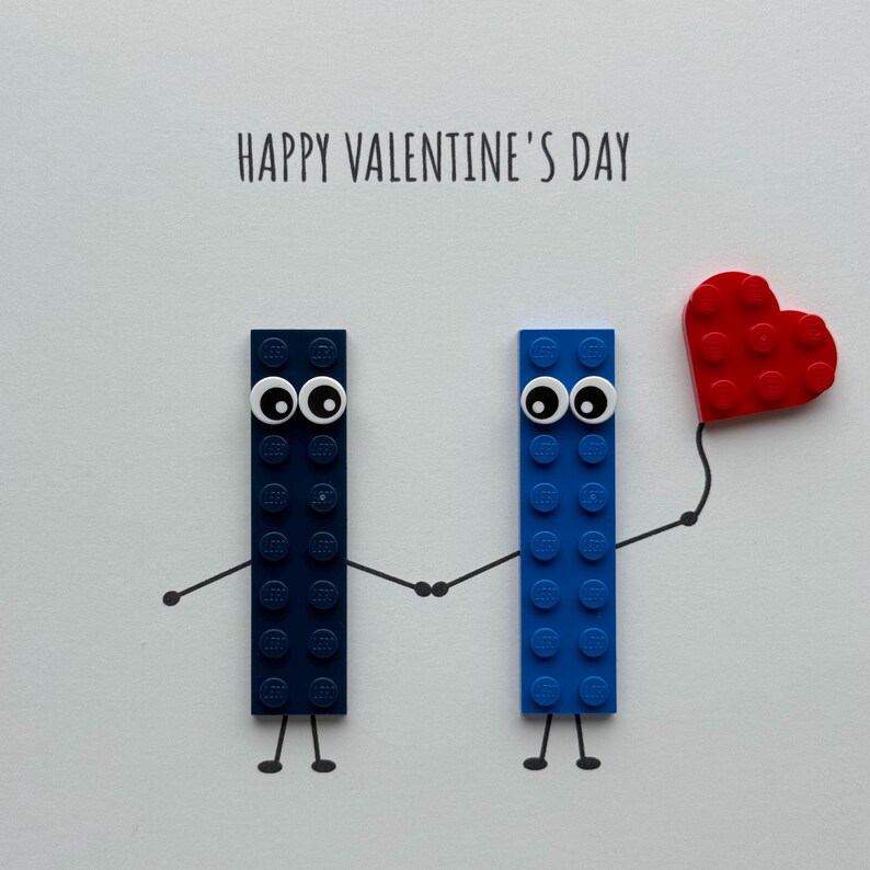 May include: A Valentine's Day card featuring two Lego brick figures with googly eyes holding hands. One figure is dark blue, the other is blue, and a red heart is held by the blue figure. The words "HAPPY VALENTINE'S DAY" are printed above.