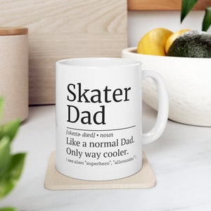 Könnte beinhalten: Eine weiße Keramiktasse mit einer schwarzen Grafik, auf der "Skater Dad" steht, und einer Definition des Begriffs. Die Definition lautet: "[skater dad] Substantiv Wie ein normaler Vater. Nur viel cooler. [siehe auch: "Superheld", "Allrounder"]"