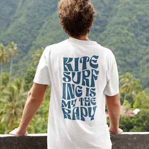 Peut inclure: T-shirt blanc avec un motif bleu qui dit "Kite Surfing is my therapy". Le t-shirt est porté par une personne debout sur un balcon surplombant une montagne verdoyante.
