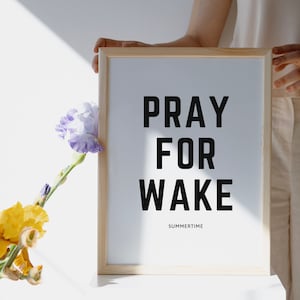 May include: A framed print with the text "Pray for Wake" and the word "Summertime" below it. The print is held by a person's hand and is in front of a white wall with a bouquet of yellow and purple flowers.