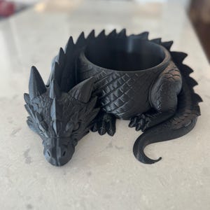 Puede incluir: Una maceta negra con forma de dragón y superficie texturizada. El dragón está tumbado con la cabeza apoyada en la superficie. La maceta tiene una abertura redonda para plantas u otros objetos. Las escamas y los detalles del dragón son claramente visibles.