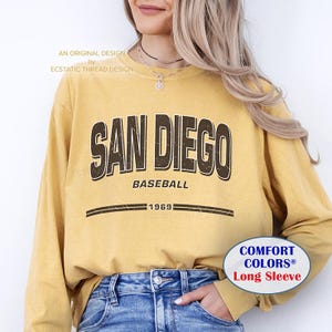 May include: A long-sleeved mustard yellow sweatshirt with the text "SAN DIEGO BASEBALL 1969" in brown lettering. The text is in a vintage style font. The sweatshirt has a comfort colors logo on the bottom right corner.