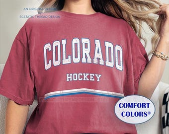 May include: A red t-shirt with a white distressed graphic that says "COLORADO HOCKEY" in a vintage style. The shirt has a blue, white, and gray stripe across the bottom and the words "COMFORT COLORS®" in a white oval on the bottom right.