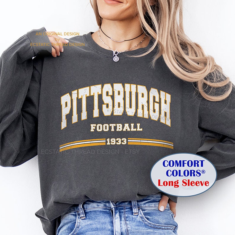 Op de afbeelding: Donkergrijs shirt met lange mouwen met het woord "PITTSBURGH" in versleten witte letters. Daaronder staat "FOOTBALL 1933" in een vergelijkbare stijl gedrukt, met gouden en witte strepen. Het shirt heeft een "COMFORT COLORS&reg; Long Sleeve" label.