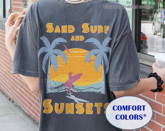 May include: A dark gray t-shirt with a vintage-style graphic of a surfer riding a wave at sunset. The graphic includes palm trees, a sunset, and the text "Sand Surf and Sunsets". The shirt also has the text "Comfort Colors®" printed on the bottom right corner.