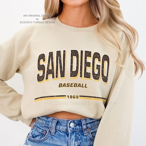 May include: A beige sweatshirt with a brown and yellow graphic that says "San Diego Baseball 1969".