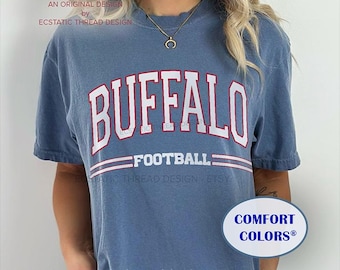 May include: A blue short-sleeved t-shirt with the word "BUFFALO" in white block letters with a red outline. Below the word "BUFFALO" is the word "FOOTBALL" in white. The shirt has the text "COMFORT COLORS®" on a white oval.