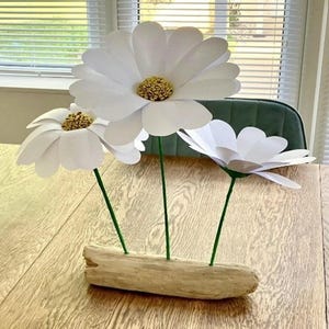 May include: Three large, handmade white paper daisies with yellow centers and green stems are arranged in a natural wood base. The flowers are displayed on a light-colored wooden table, creating a simple, elegant floral arrangement.