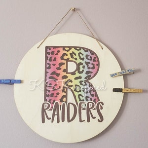 Può includere: Un cartello rotondo in legno con una lettera "R" con stampa leopardata e la parola "RAIDERS" in marrone. La lettera è riempita di colori arcobaleno. Tre mollette sono attaccate al cartello.
