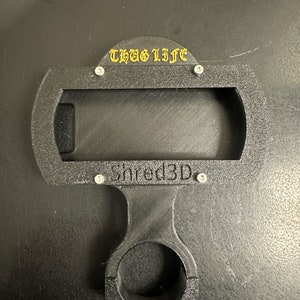 Peut inclure: Support de t&eacute;l&eacute;phone noir imprim&eacute; en 3D avec un collier de guidon noir. Le support a une d&eacute;coupe rectangulaire pour un t&eacute;l&eacute;phone et le texte "THUG LIFE" et "Shred3D" imprim&eacute; dessus.