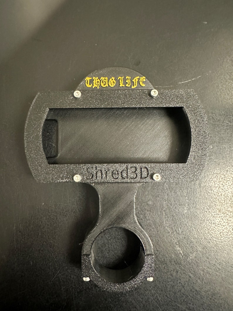 Peut inclure: Support de t&eacute;l&eacute;phone noir imprim&eacute; en 3D avec un collier de guidon noir. Le support a une d&eacute;coupe rectangulaire pour un t&eacute;l&eacute;phone et le texte "THUG LIFE" et "Shred3D" imprim&eacute; dessus.