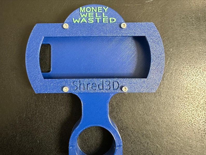 Peut inclure: Un support de t&eacute;l&eacute;phone imprim&eacute; en 3D bleu avec le texte "MONEY WELL WASTED" en haut et "Shred3D" en bas.