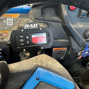 Peut inclure: Un indicateur noir avec un affichage num&eacute;rique rouge indiquant "71%" et le texte "READING ECU". L'indicateur est mont&eacute; sur un panneau noir avec le texte "BAD Side x Sides" et "Shred3D".