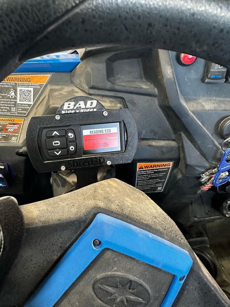 Peut inclure: Un indicateur noir avec un affichage num&eacute;rique rouge indiquant "71%" et le texte "READING ECU". L'indicateur est mont&eacute; sur un panneau noir avec le texte "BAD Side x Sides" et "Shred3D".