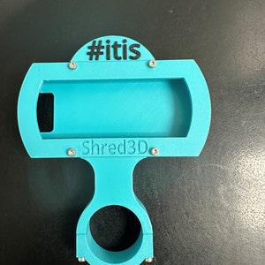 Peut inclure: Un support de t&eacute;l&eacute;phone imprim&eacute; en 3D bleu turquoise avec un collier de serrage pour guidon. Le support a une fente pour un t&eacute;l&eacute;phone et le texte "#itis" et "Shred3D" imprim&eacute; dessus.